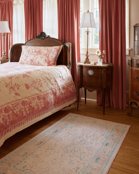 A Victorian bedroom with pink drapes, pink and white bedding, and a light pink and blue printed rug