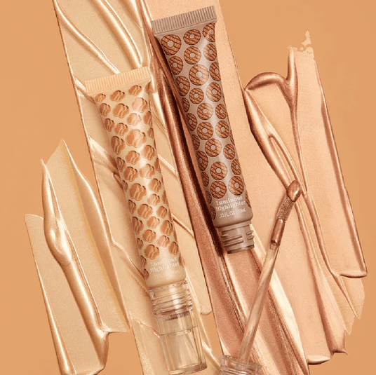 Two tubes of Hard Candy x Girl Scouts. Cookie Icing Face Highlighter laid next to each other over several highlighter swatches