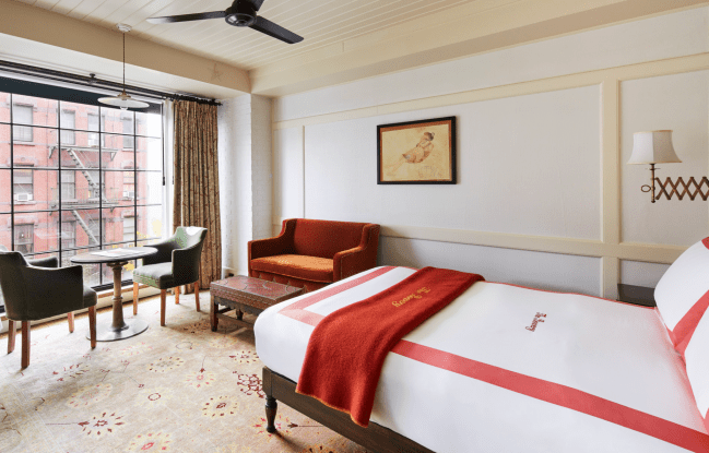A bedroom at The Bowery Hotel, featuring red-trim bedding, a small dining nook, and large city-view windows