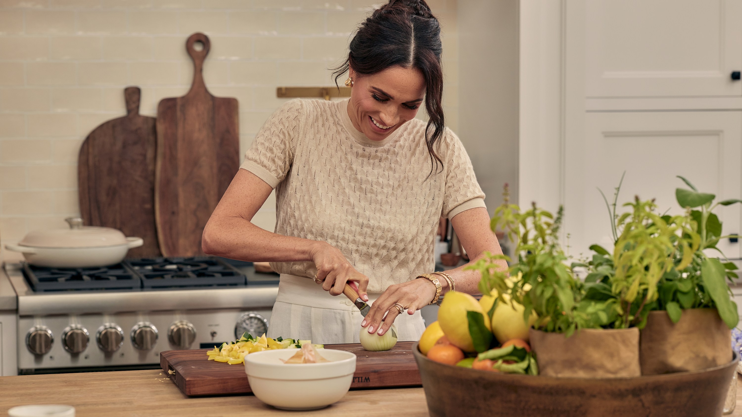 Meghan Markle in a kitchen, cutting an onion on a wooden cutting board.