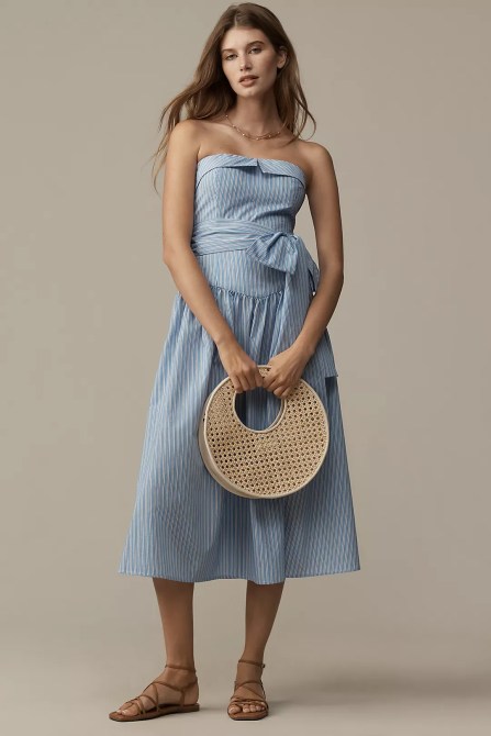 A woman wearing a blue strapless dress, holding a rattan bag.
