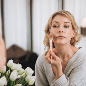 Side view of a beautiful adult blonde woman applying blush while looking into a mirror in her cozy bedroom. Morning skincare, cosmetics, and self-care routine