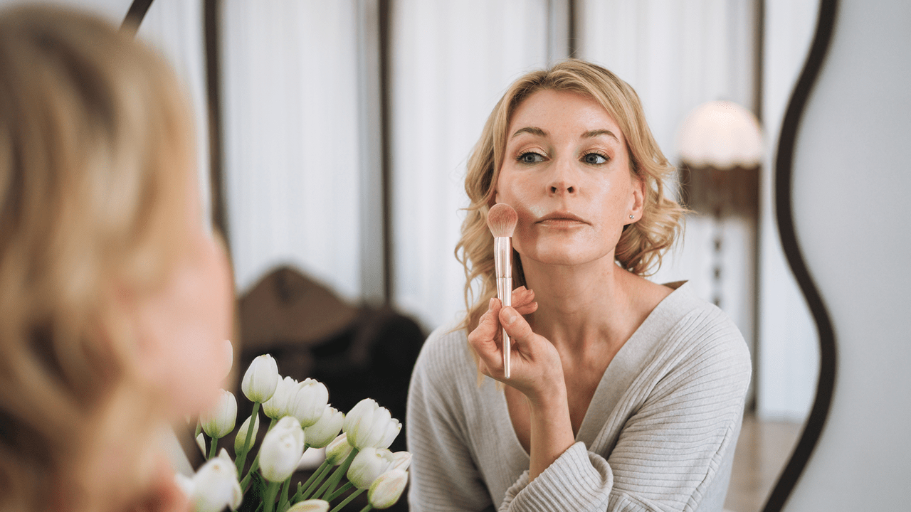 Side view of a beautiful adult blonde woman applying blush while looking into a mirror in her cozy bedroom. Morning skincare, cosmetics, and self-care routine